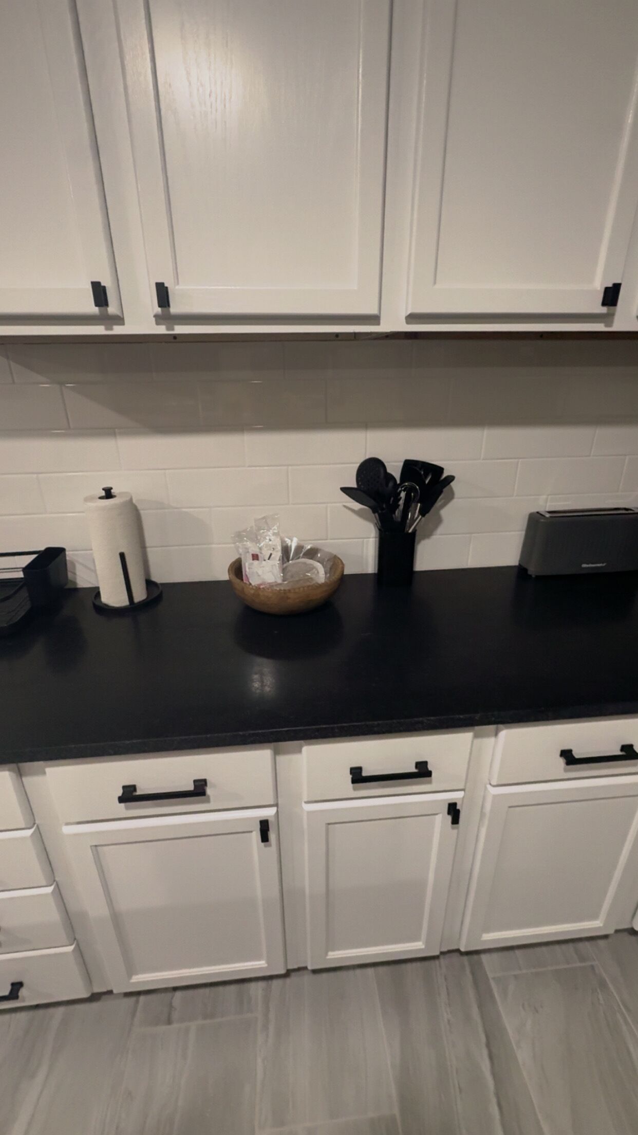 Eine Küche mit weißen Schränken, einer schwarzen Arbeitsplatte, einem Toaster, einem Papierhandtuchhalter, einer Schüssel mit Besteck und einem Mülleimer.