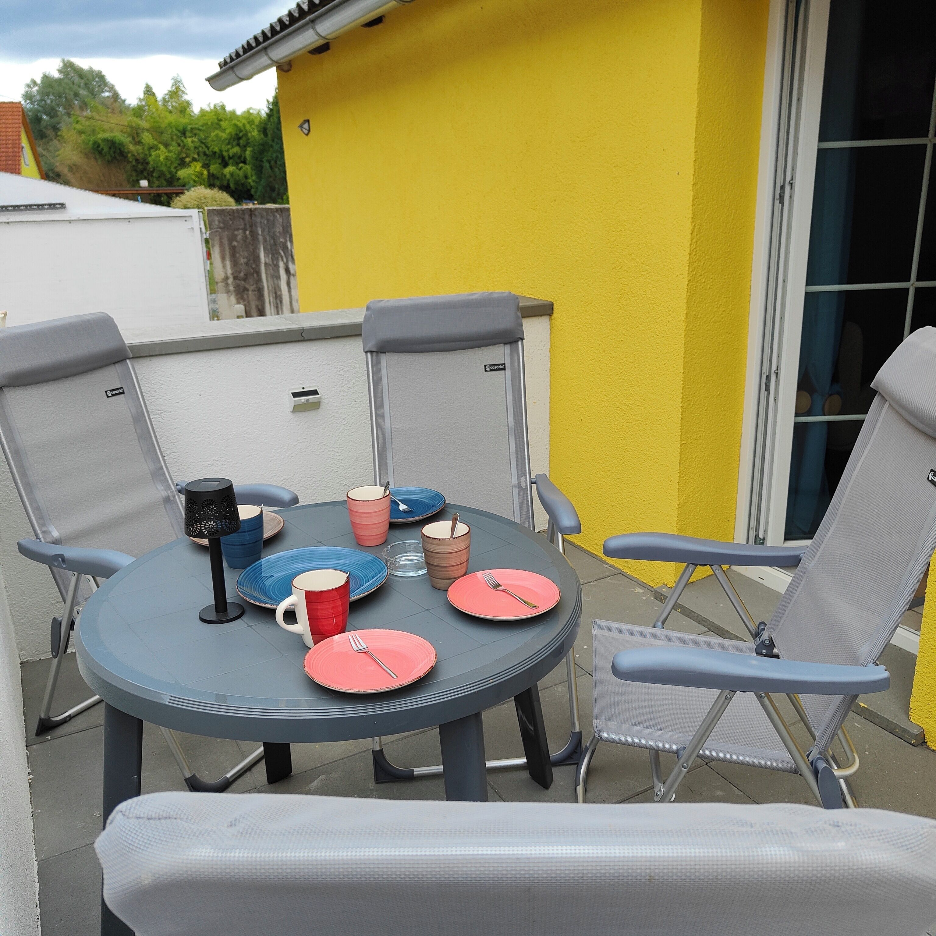 An outdoor dining area with a table and four chairs, a yellow wall, and a window.