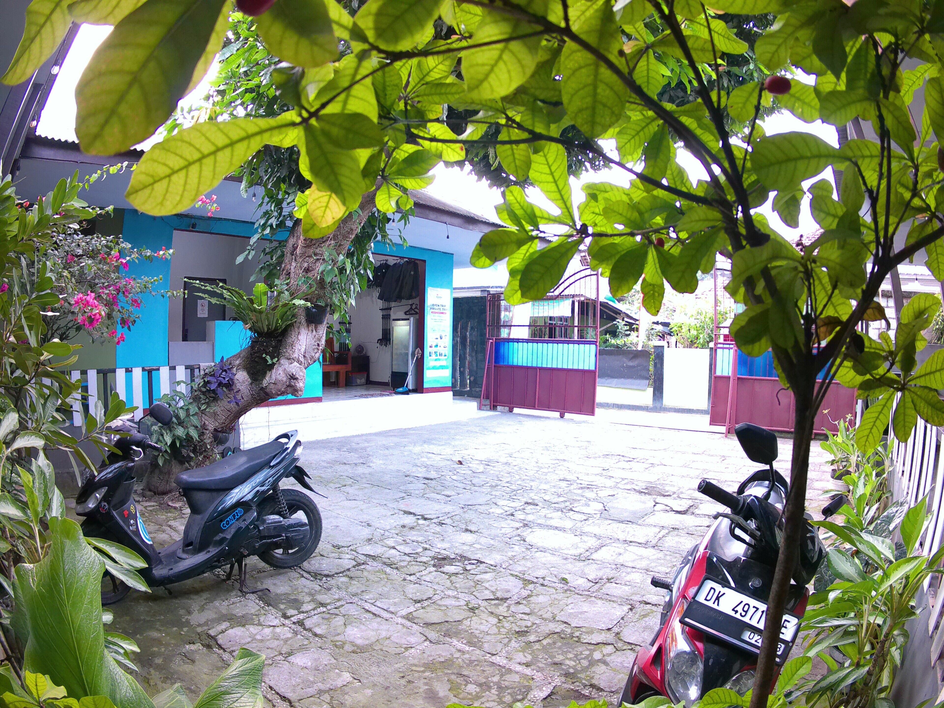 Um pátio com uma scooter estacionada na sombra, uma motocicleta em primeiro plano e um edifício com fachada azul ao fundo.