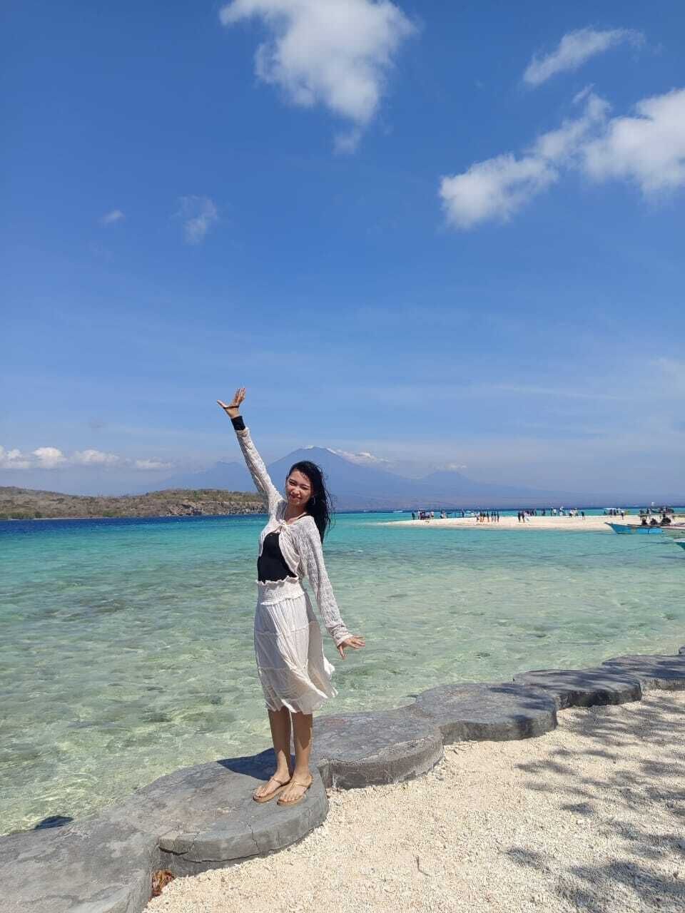 Una persona está de pie sobre un camino de piedra frente a un mar azul y transparente, con una montaña y una playa a lo lejos.