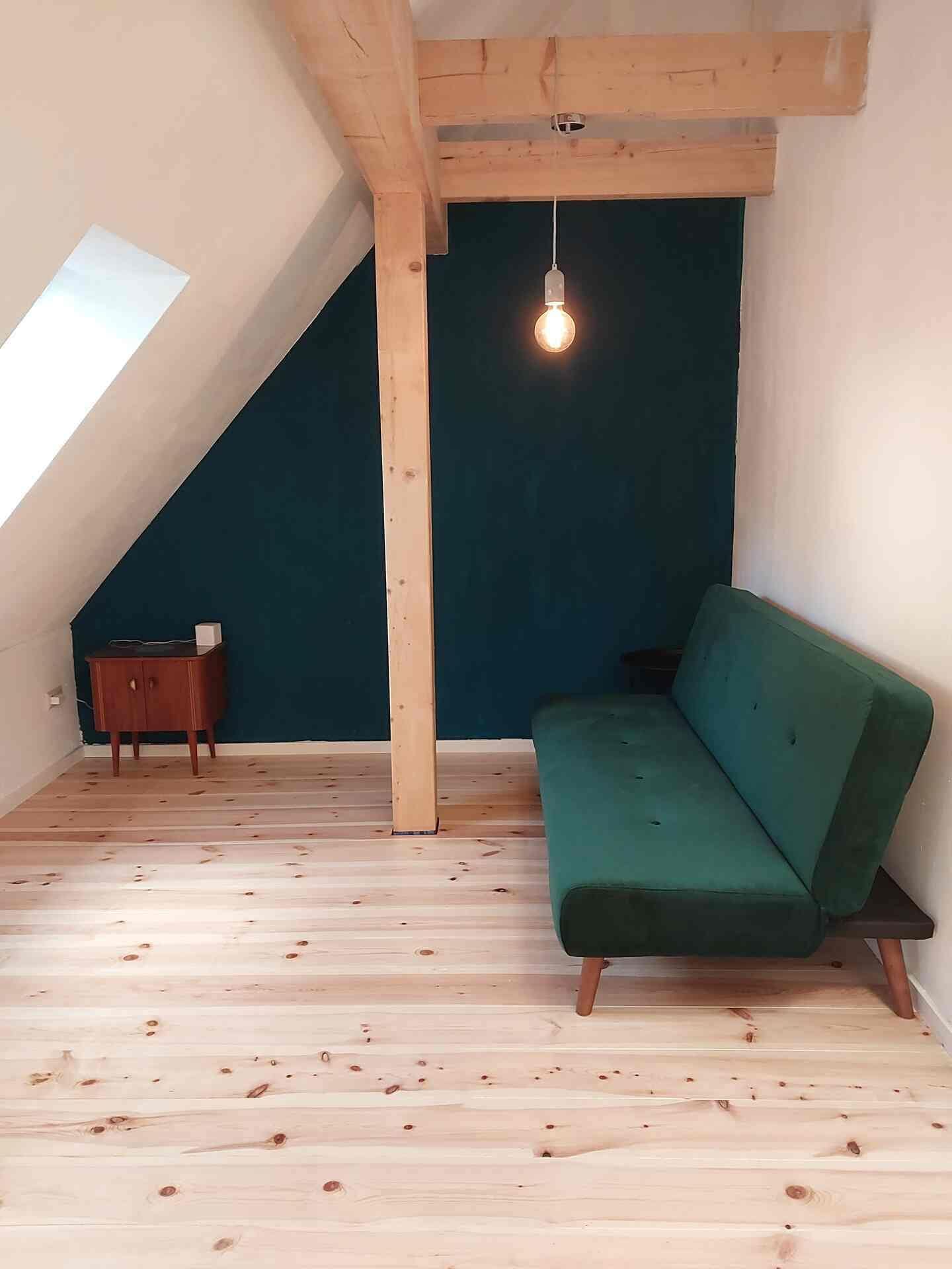 Ein Zimmer mit schrägem Dach, einem grünen Sofa, einer kleinen Holzkabine und einer hängenden Glühbirne.