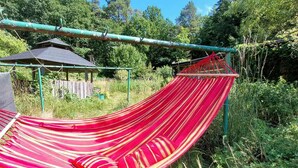 En hængekøje spændt ud mellem to stænger i et naturområde med træer og en træbygning i baggrunden.