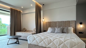 A modern bedroom with a large bed, a sofa, a small table, and a view of greenery outside. - Stella Elite Suites (Zakynthos)