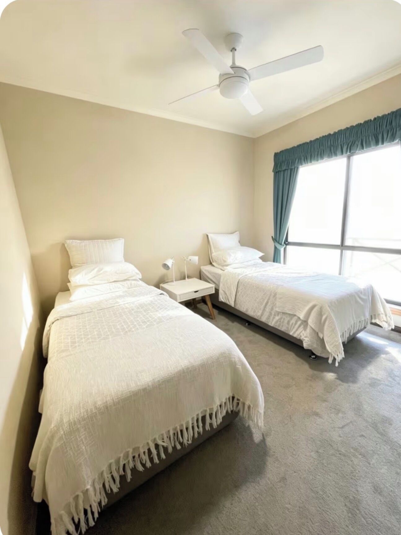 A hotel room with two beds, a ceiling fan, a window with curtains, and a nightstand with a lamp.
