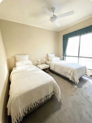 Ein Hotelzimmer mit zwei Betten, einem Deckenventilator, einem Fenster mit Vorhängen und einem Nachttisch mit Lampe.