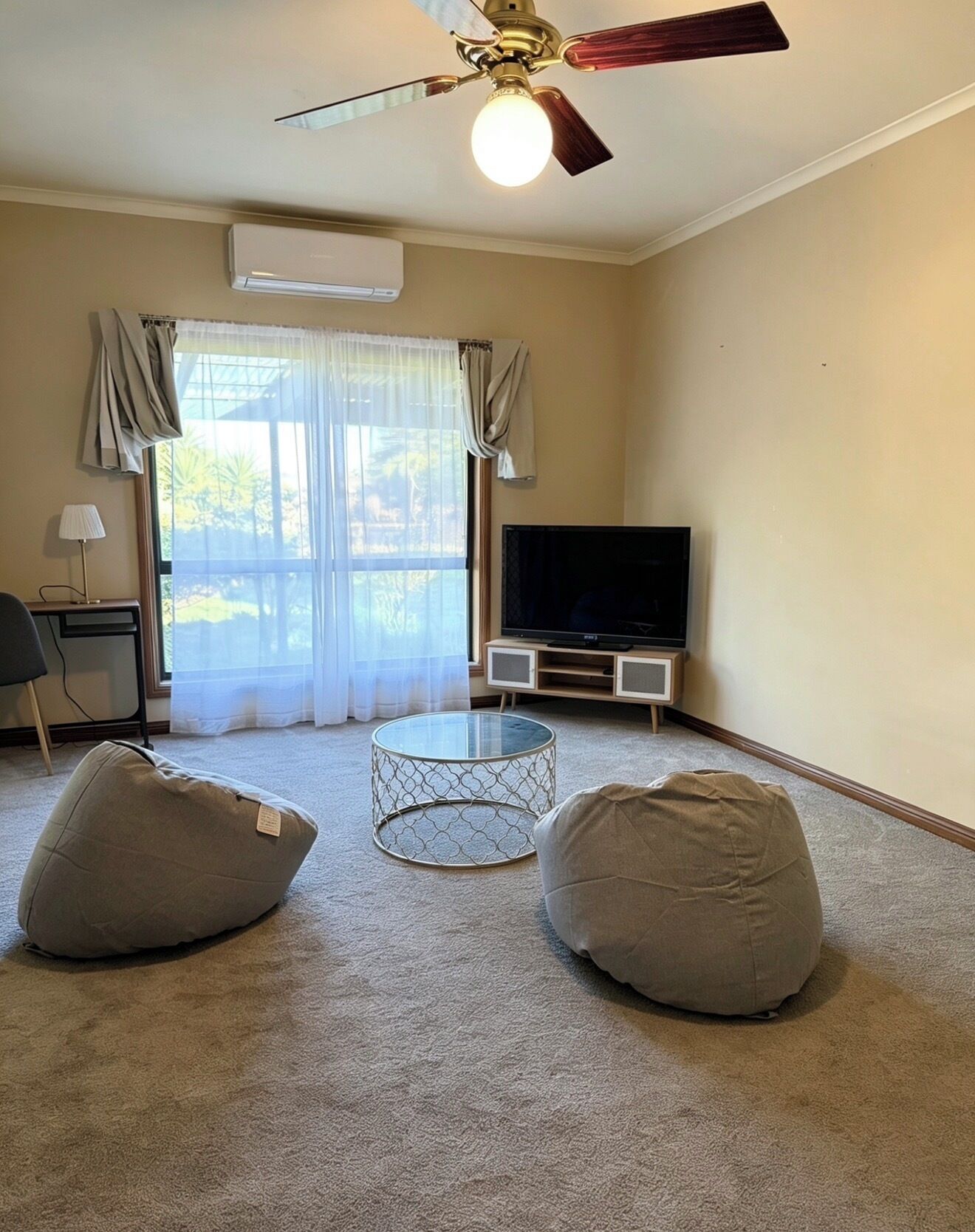 A living room with a ceiling fan, a flat-screen TV, two bean bags, a glass coffee table, and a window with curtains.