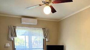 Un salon doté d’un ventilateur de plafond, d’un téléviseur à écran plat, de deux fauteuils poire, d’une table basse en verre et d’une fenêtre avec des rideaux.