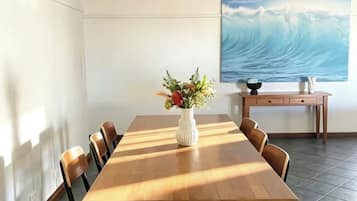 A dining area with a long wooden table, six chairs, a vase with flowers, and a painting of a wave on the wall.