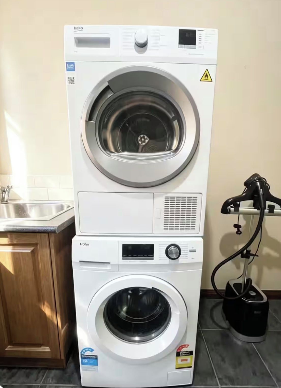 A stacked washer and dryer unit with a control panel, a warning label, and a hose connected to the dryer.