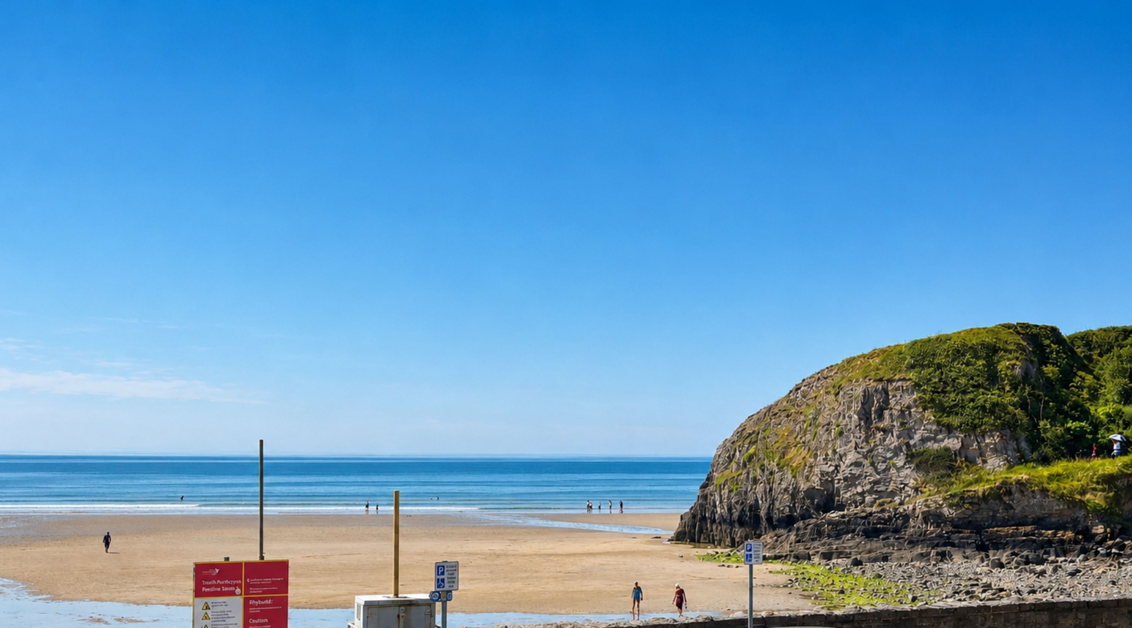 A beach with a rocky cliff, clear blue sky, and a few people walking.