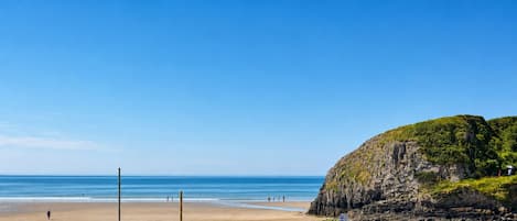 A beach with a rocky cliff, clear blue sky, and a few people walking.