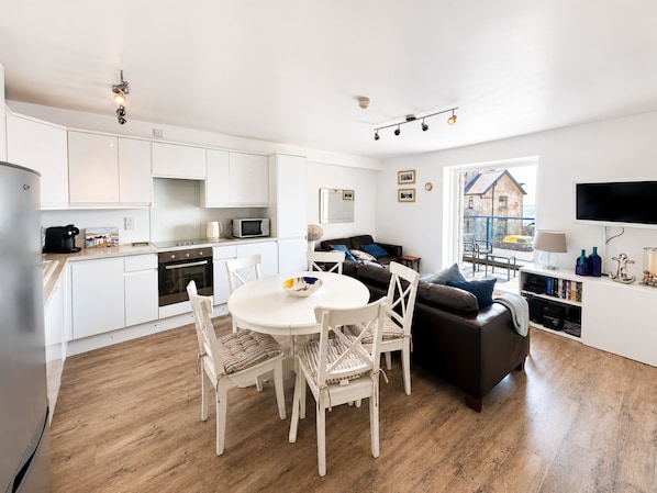 A modern kitchen with white cabinets, a dining area with a round table and chairs, a black sofa, and a television on a white stand.