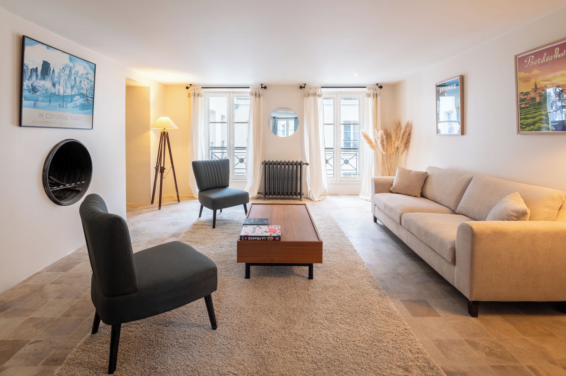 A living room with a sofa, armchair, coffee table, and a radiator. There are windows with curtains, a lamp, and artwork on the walls.