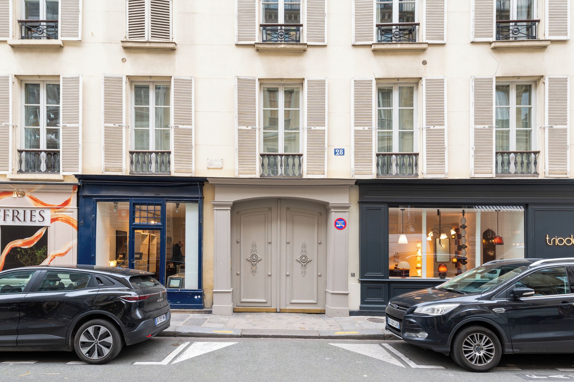 A street view with two cars parked on the side, a building with multiple windows and shutters, and a door with a sign above it.