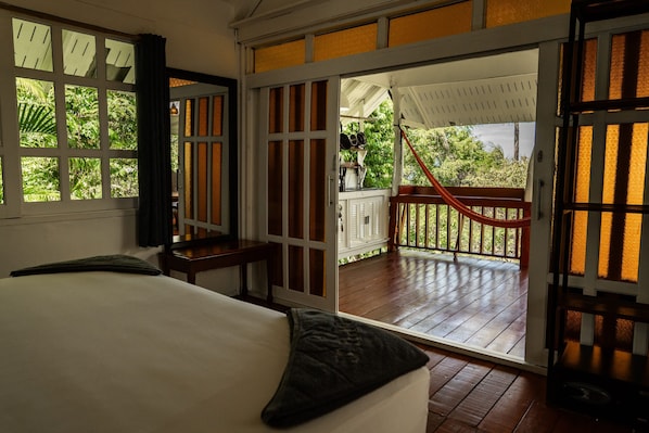A room with a bed, a window with a view of greenery, and a door leading to an outdoor area with a hammock.