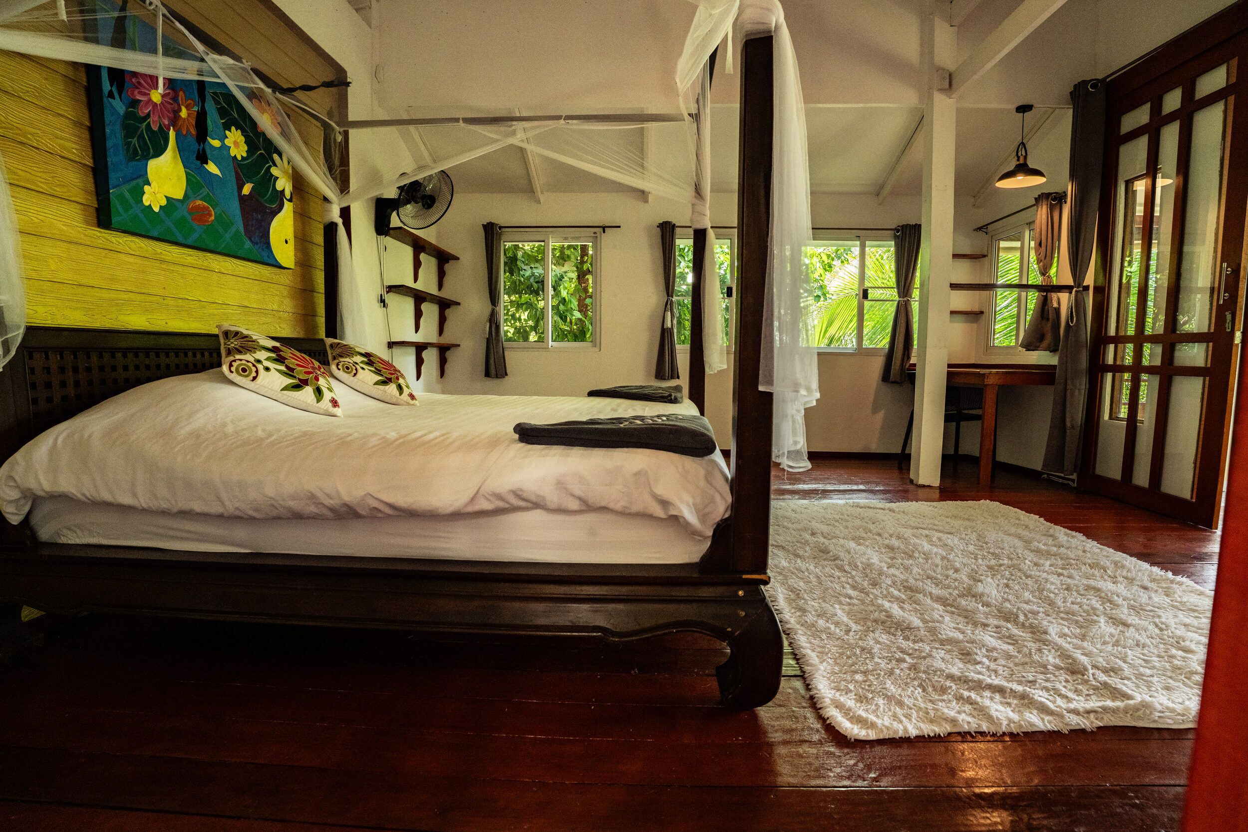 A four-poster bed with white linens and decorative pillows, a wooden floor, and a large window with curtains.