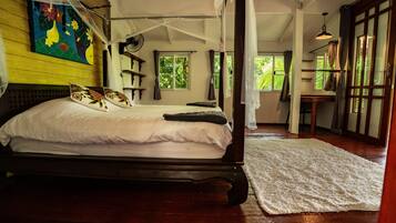 A four-poster bed with white linens and decorative pillows, a wooden floor, and a large window with curtains.