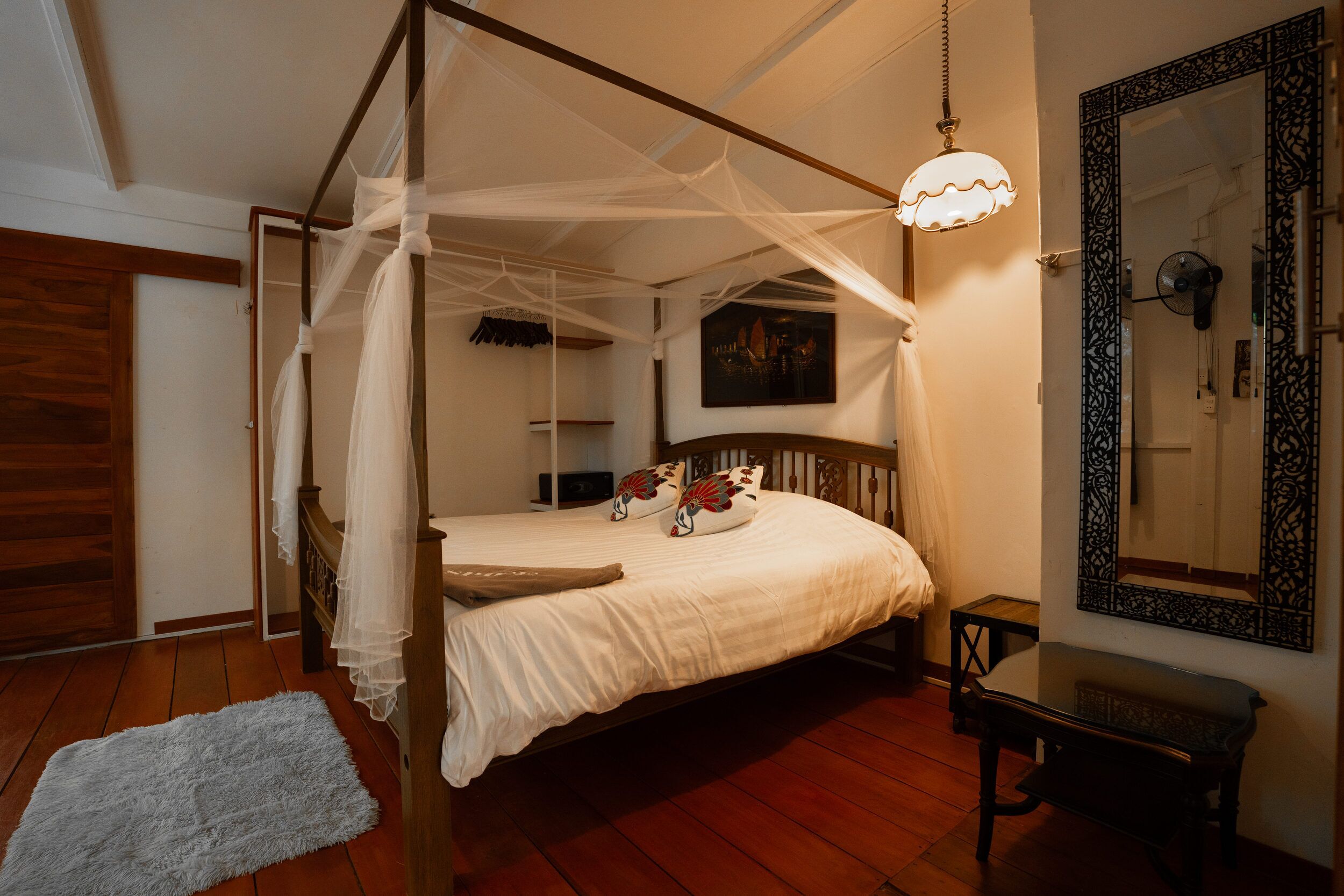 Ein Bett mit Baldachin, weiße Bettdecke, Nachttisch, Wandventilator und ein dekorativer Spiegel.
