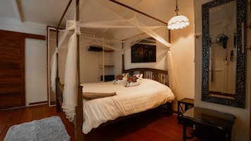 Una cama con dosel y cuatro columnas, con una colcha blanca, una mesita de noche, un ventilador de pared y un espejo decorativo.