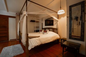 Una cama con dosel y cuatro columnas, con una colcha blanca, una mesita de noche, un ventilador de pared y un espejo decorativo.