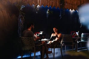 Two people seated at a table with a laptop, in a room with patterned chairs and a thatched ceiling.