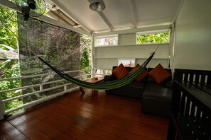Una hamaca en una habitación con piso de madera, un sofá con cojines y vista a zonas verdes al exterior.