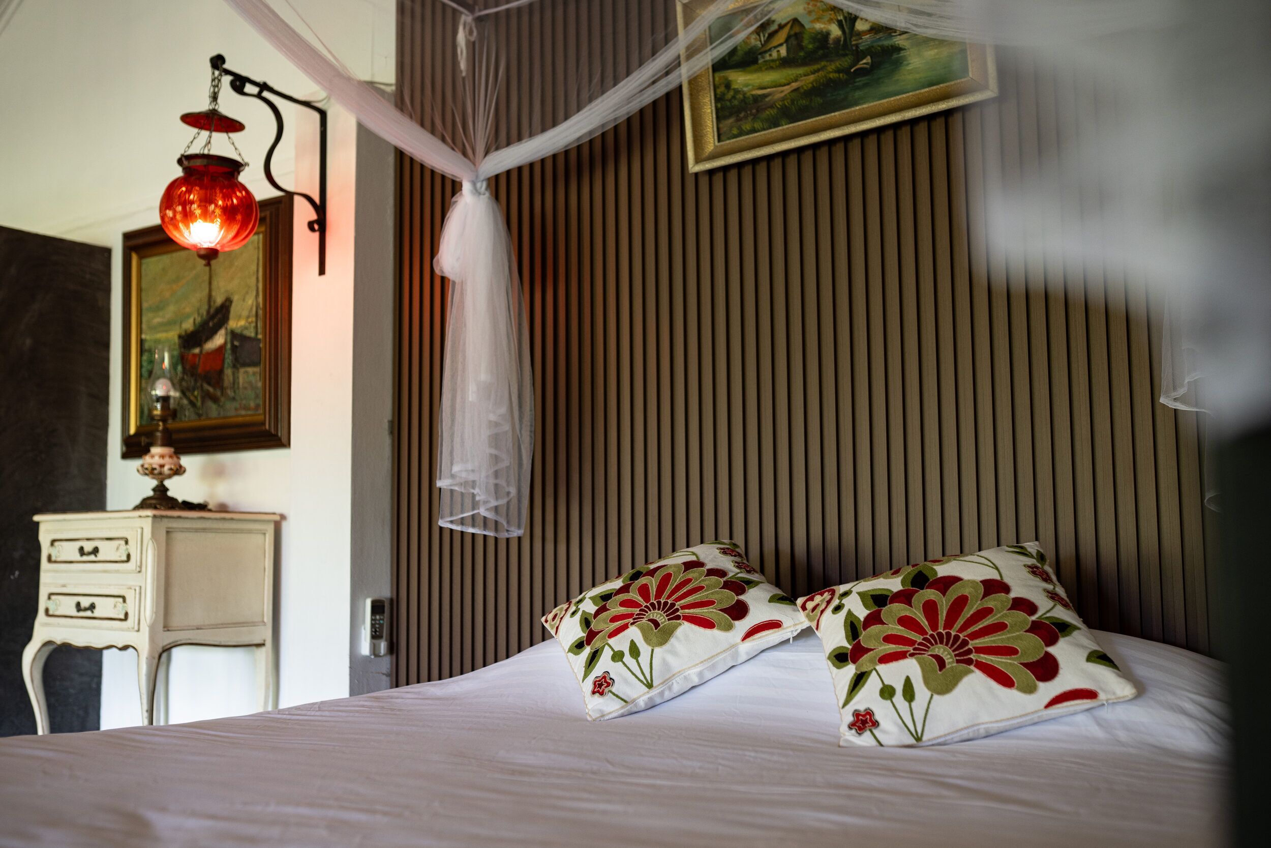 A bedroom with a bed, pillows, a nightstand, a lamp, a painting, and a window with blinds.