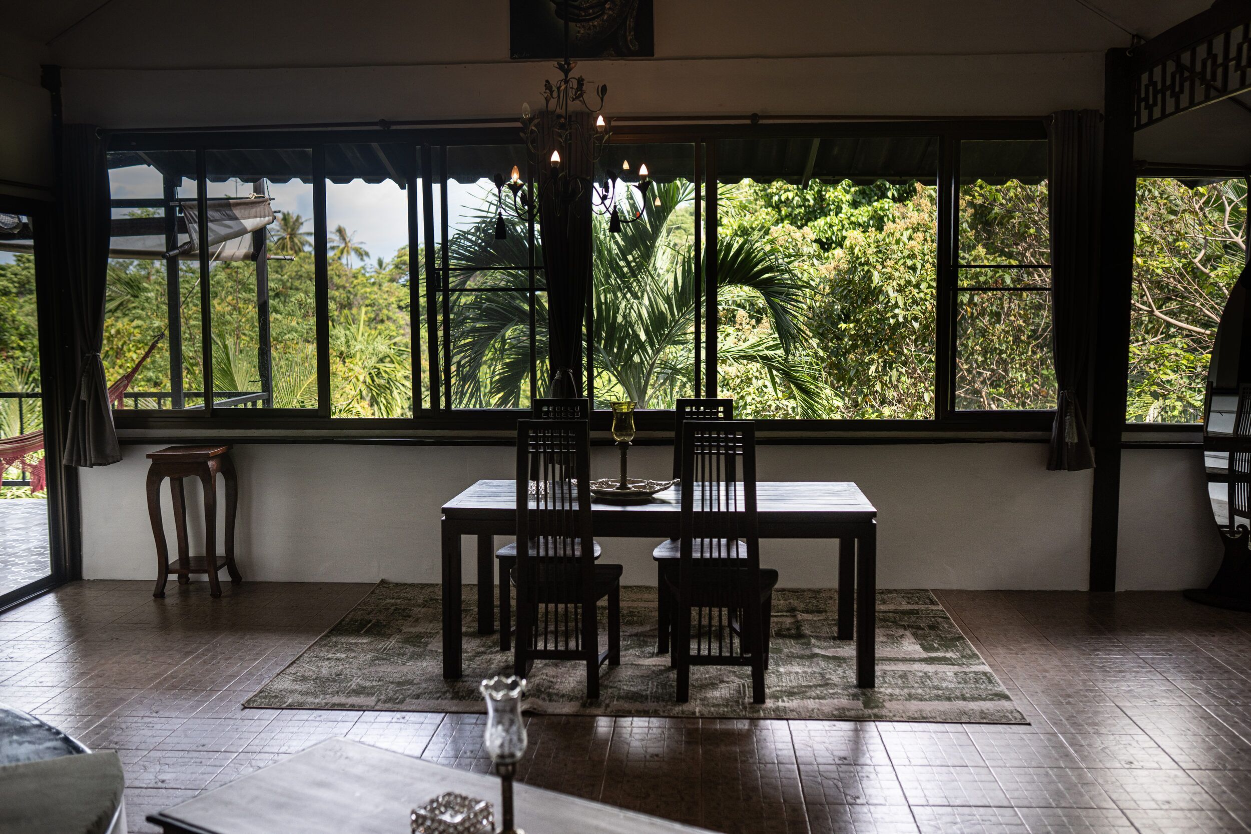 Uma área de refeições com mesa e cadeiras, janelas amplas com vista para o verde e um lustre.
