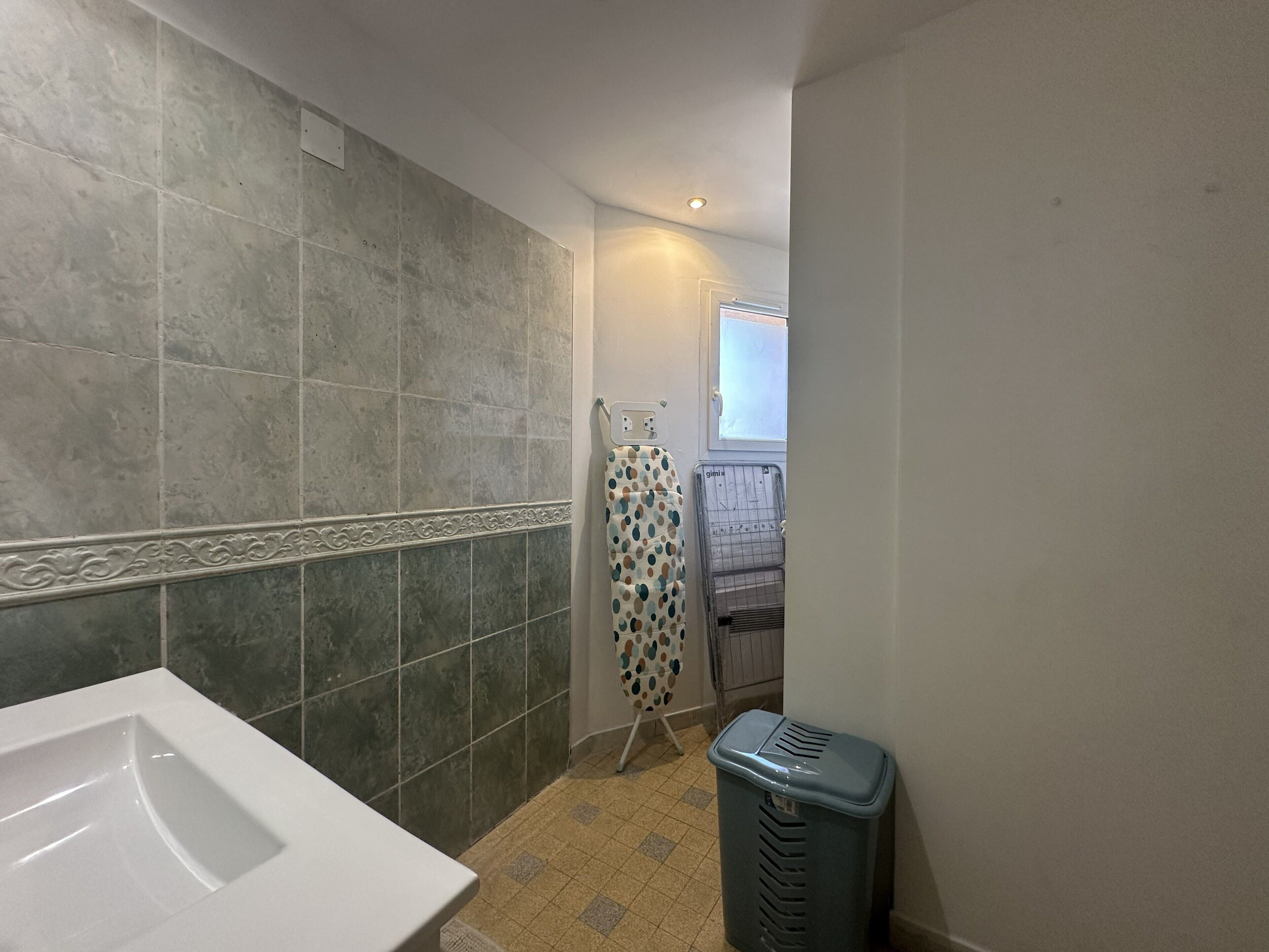 Une salle de bain avec un lavabo blanc, des murs carrelés, un rideau de douche à motifs et une poubelle.