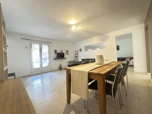 En rummelig opholdsstue med spisebord, stole, et fjernsyn og et stort vindue.