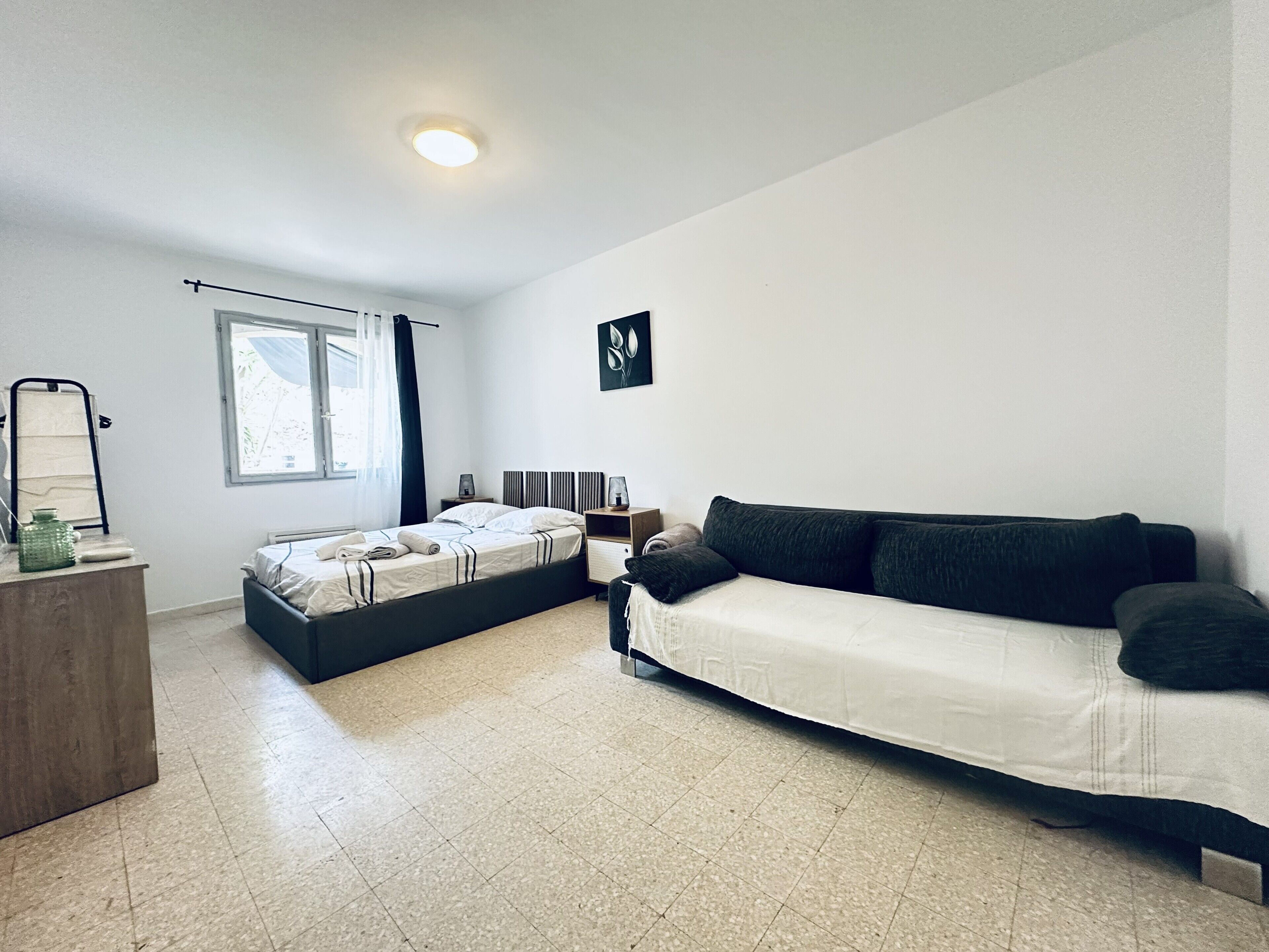 A modern bedroom with a bed, a sofa, a bedside table, a mirror, and a window.