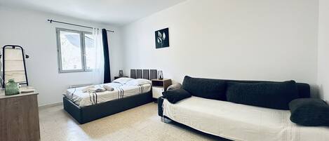 A modern bedroom with a bed, a sofa, a bedside table, a mirror, and a window.