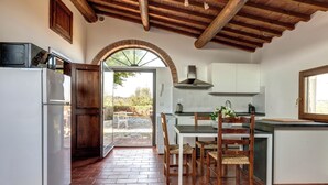 A kitchen with a brick floor, white cabinets, a wooden dining table with chairs, and a view through an arched doorway to an outdoor area. - Le Mortinete Farmstay: Colombino (Toscana)