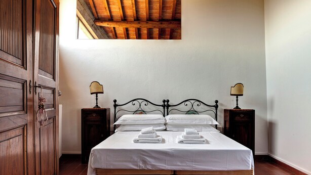 A neatly made bed with white linens and two bedside tables with lamps, in a room with wooden flooring and a wooden door. - Le Mortinete Farmstay: Colombino (Toscana)