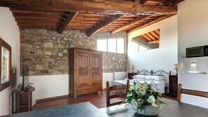 A rustic room with a stone wall, wooden ceiling beams, a bed with white linens, a wooden cabinet, and a kitchen area with a microwave. - Le Mortinete Farmstay: Colombino (Toscana)