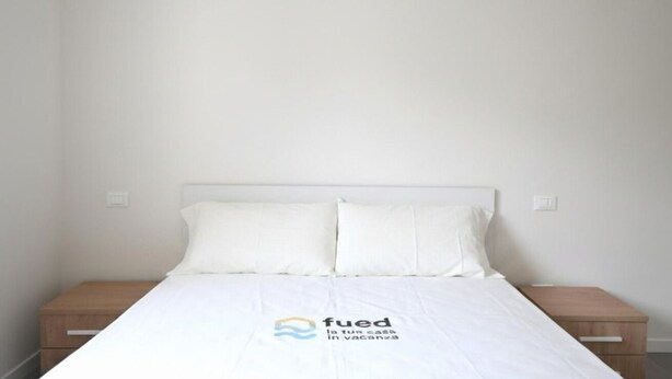 Una cama individual con colchón y almohadas blancas, una mesita de noche de madera y un interruptor de luz en la pared.