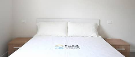 A single bed with a white mattress and pillows, a wooden nightstand, and a wall-mounted light switch.