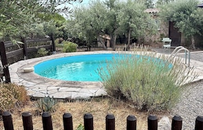 Ein runder Swimmingpool mit Steinrand, umgeben von einem Holenzaun und verschiedenen Pflanzen.