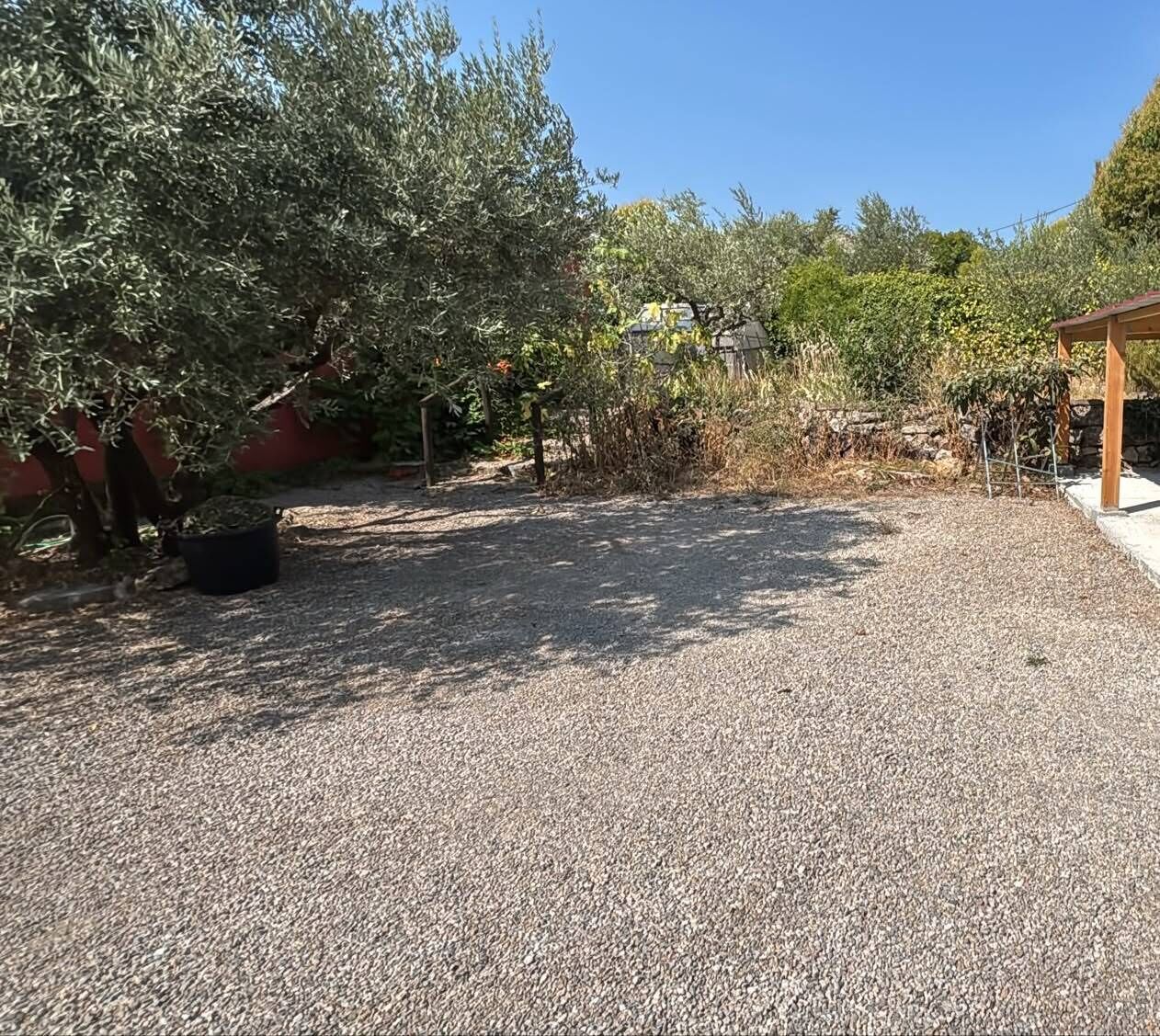 Eine Schotterterrasse mit einer Pergola, umgeben von Olivenbäumen und einer Steinmauer.