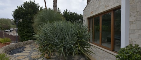 Une maison avec une façade en pierre, une grande fenêtre et un jardin où poussent différentes espèces de plantes.