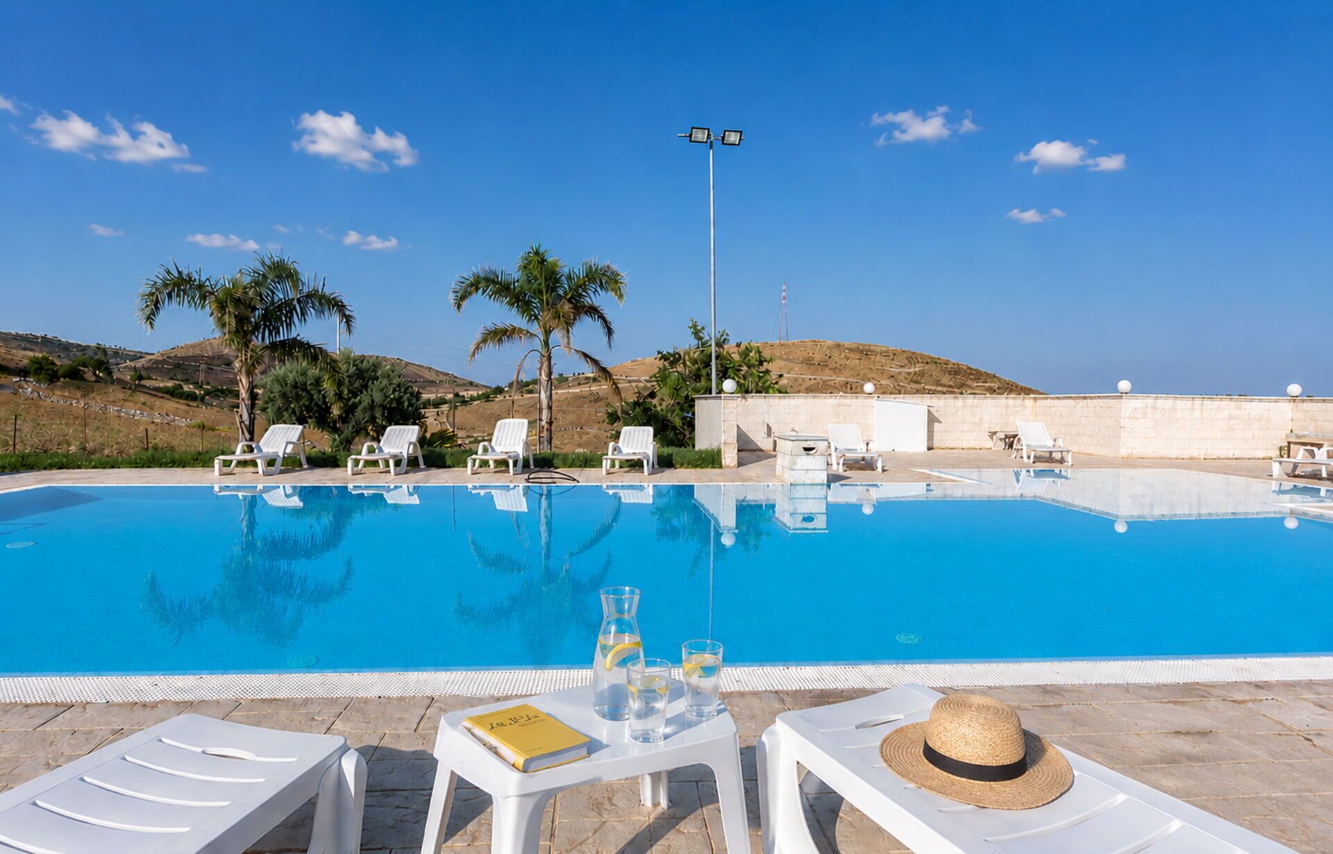 A clear blue swimming pool with white lounge chairs and a table with drinks, surrounded by palm trees and a clear sky.