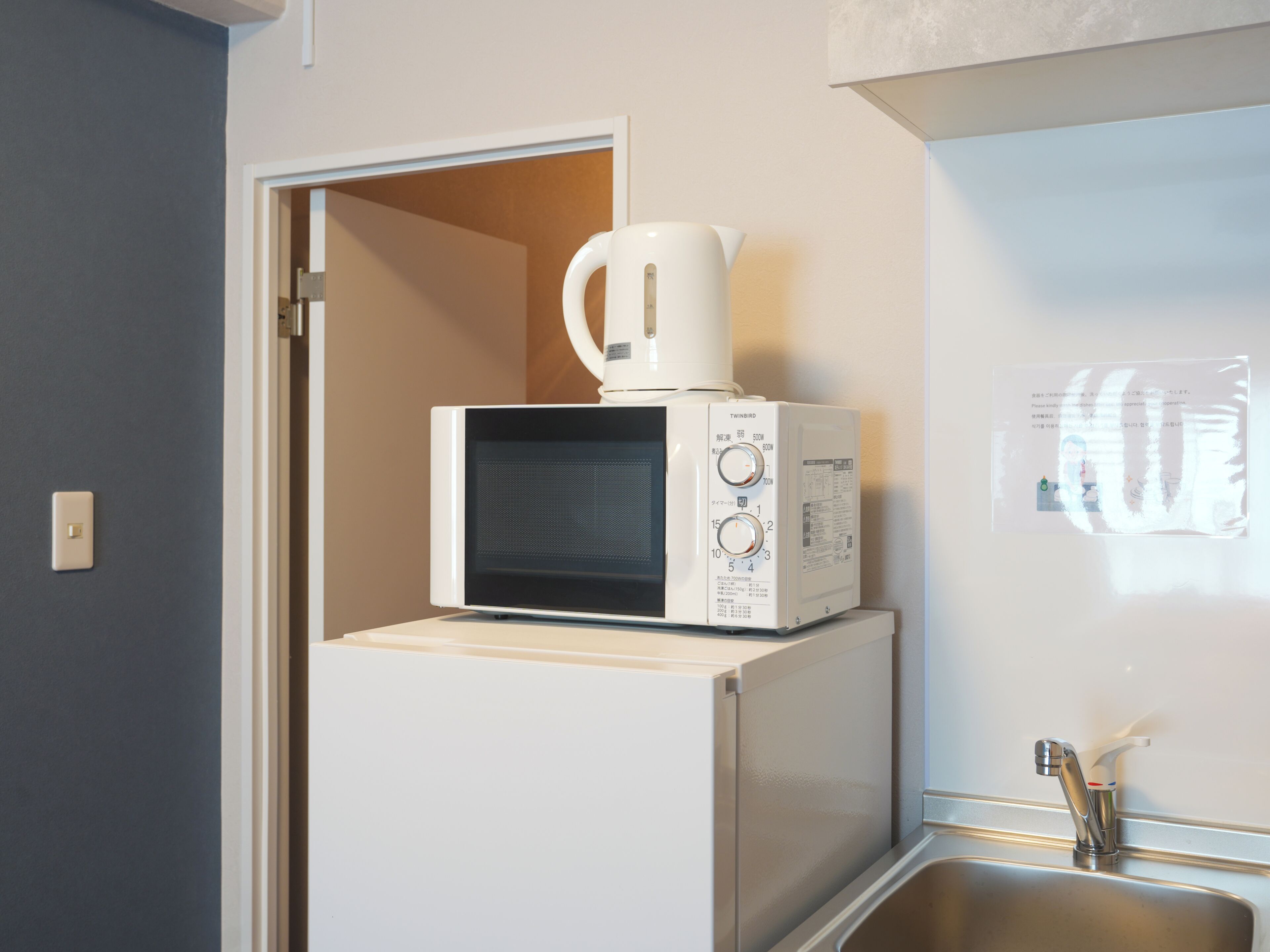 A kitchen with a microwave and a coffee maker on top of a refrigerator, a sink, and a door leading to another room.