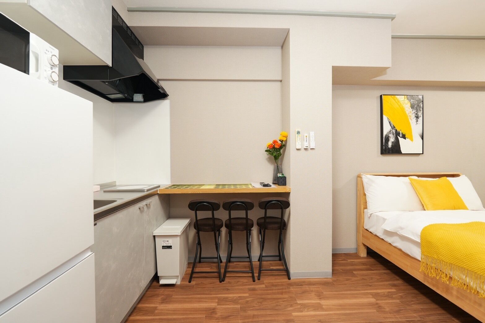 A compact room with a kitchenette, a dining area with chairs, a bed with a yellow blanket, and a painting on the wall.