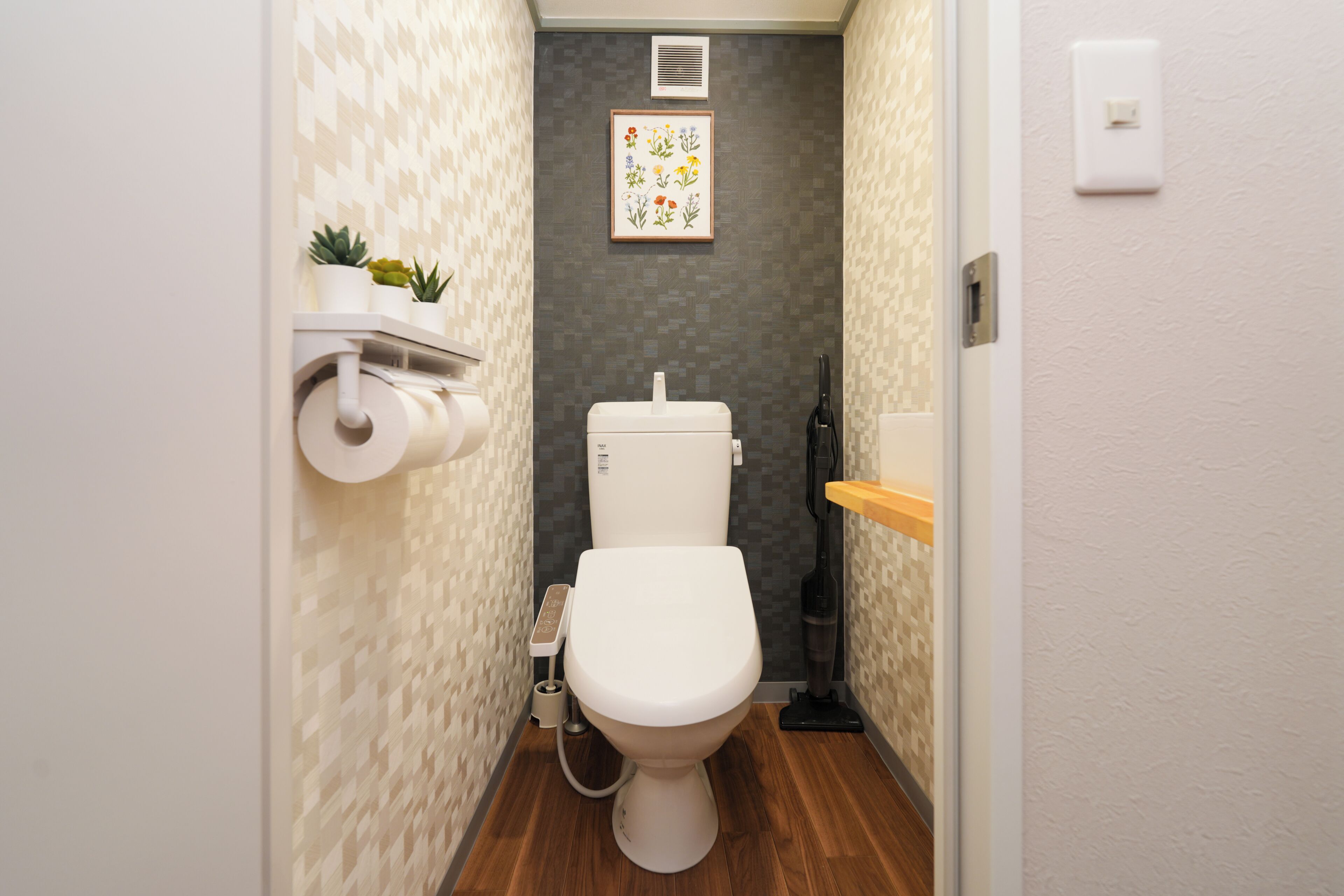A compact bathroom with a toilet, a toilet paper holder, a hand dryer, and a small shelf with plants.