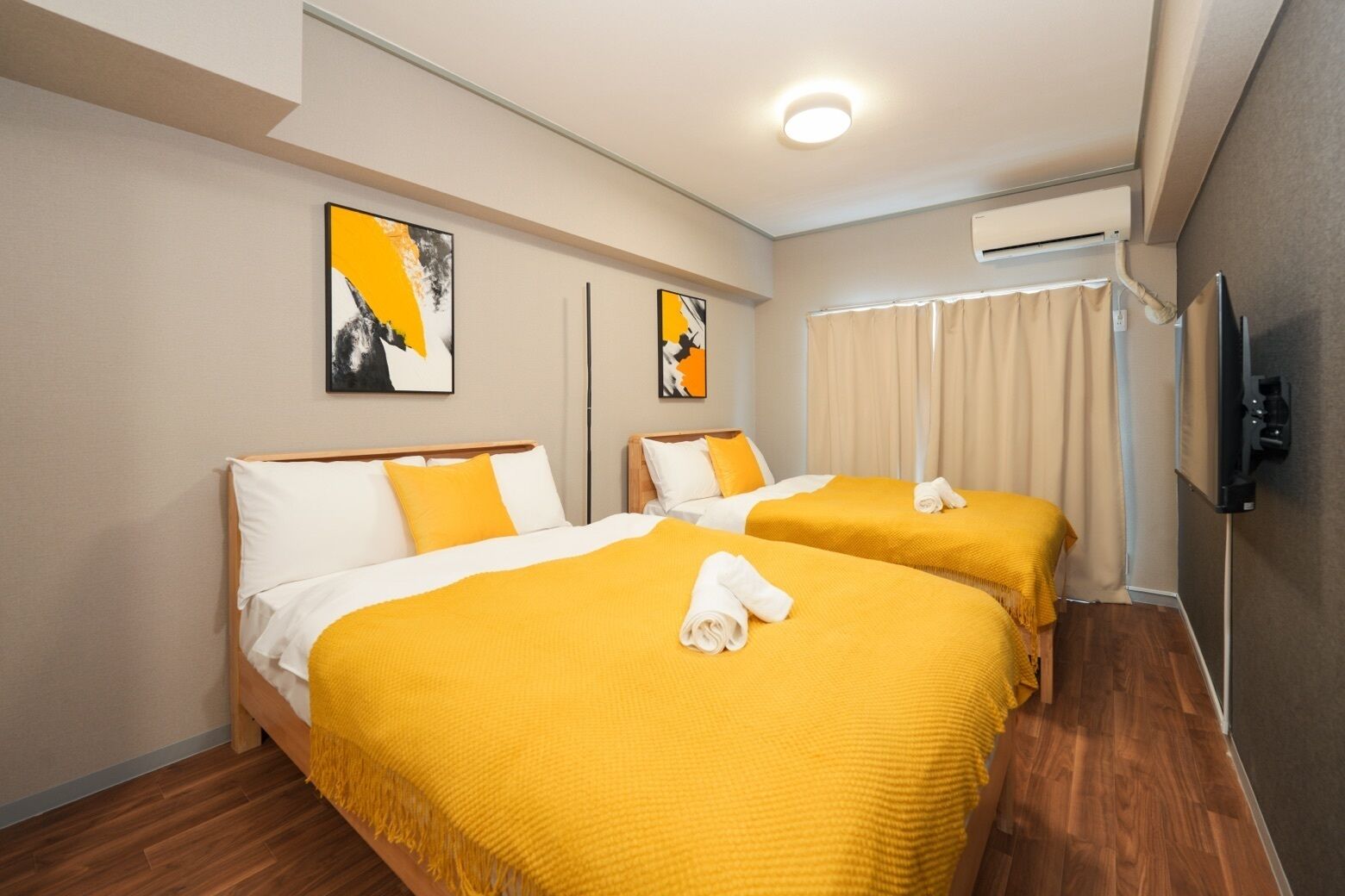 A hotel room with two beds, each with a yellow bedspread and white pillows, a flat-screen TV mounted on the wall, and artwork above the beds.