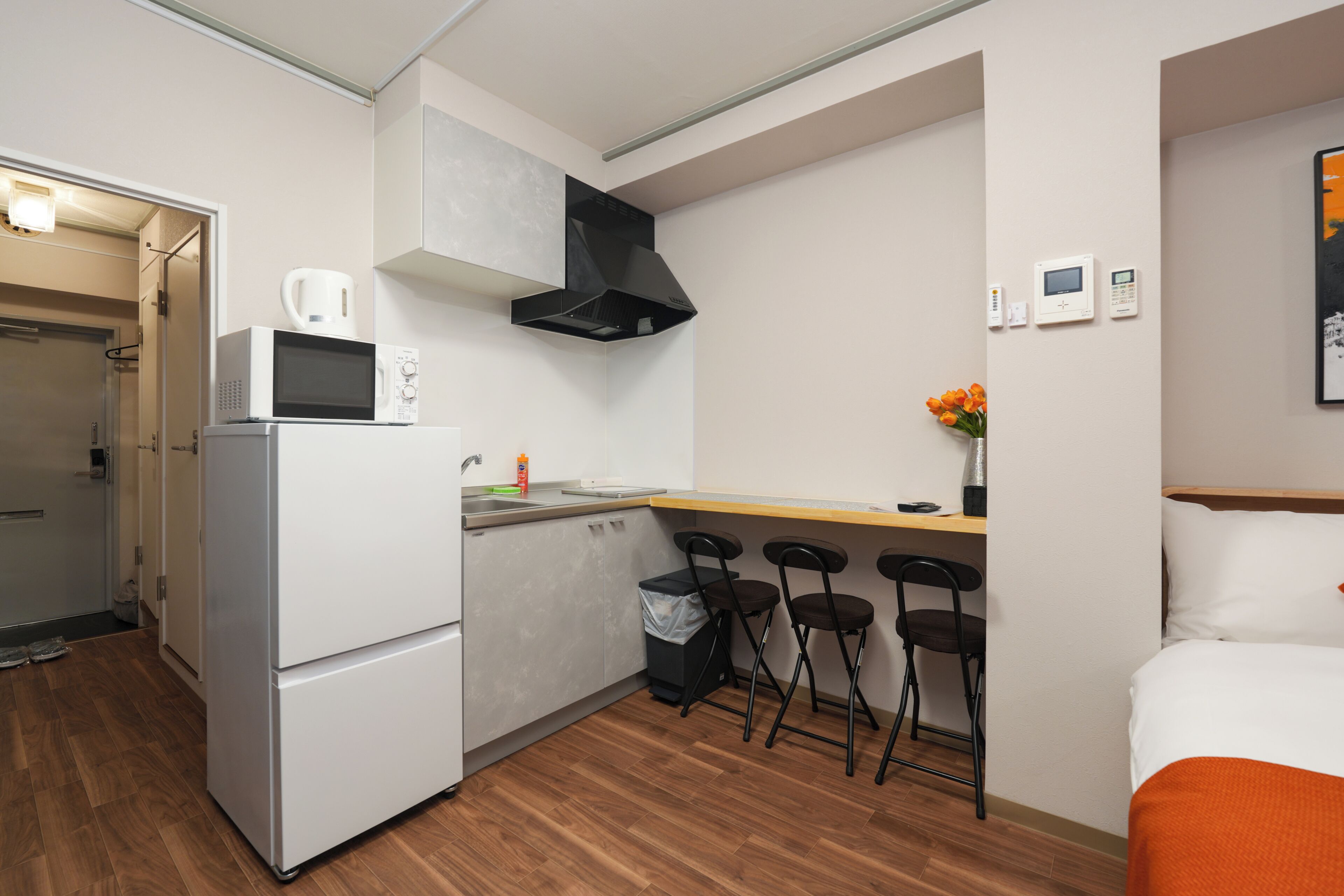 A compact kitchen with a microwave, kettle, and a small sink. There are three foldable chairs and a bed with an orange blanket.