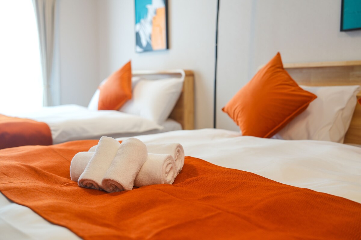 A hotel room with two beds, each with white bedding and orange pillows. There are rolled towels on the bed in the foreground.