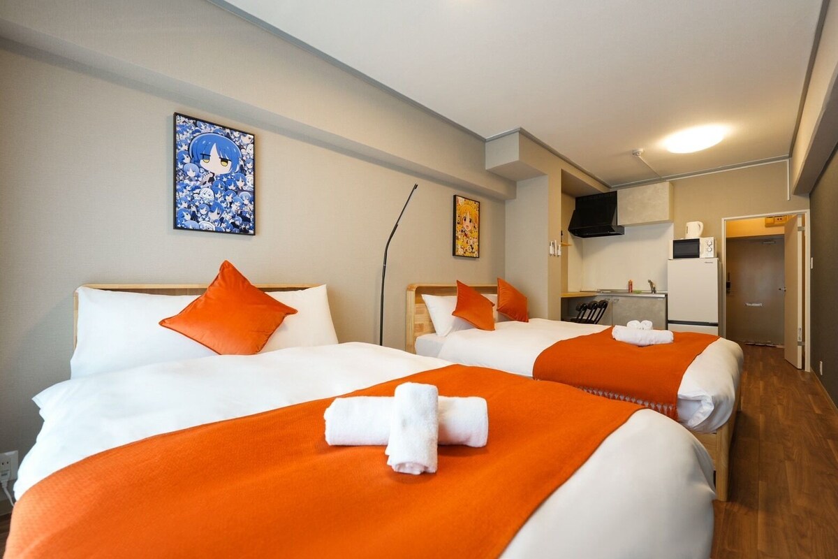A hotel room with two beds, each with white linens and orange accents, a kitchenette, and a bathroom visible through an open door.