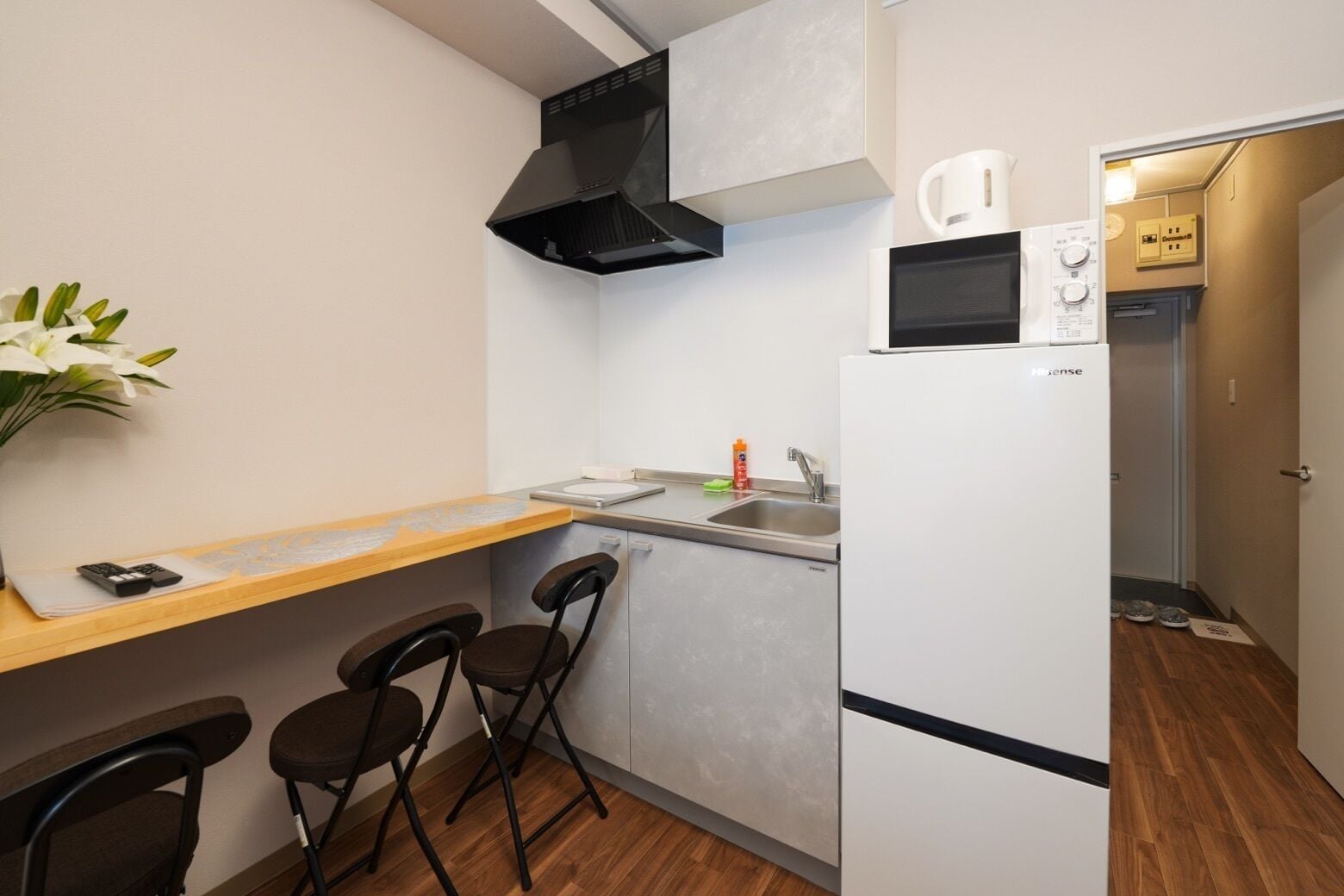 A compact kitchenette with a sink, a coffee maker, and a refrigerator. There are two stools and a small table with a remote control on it.