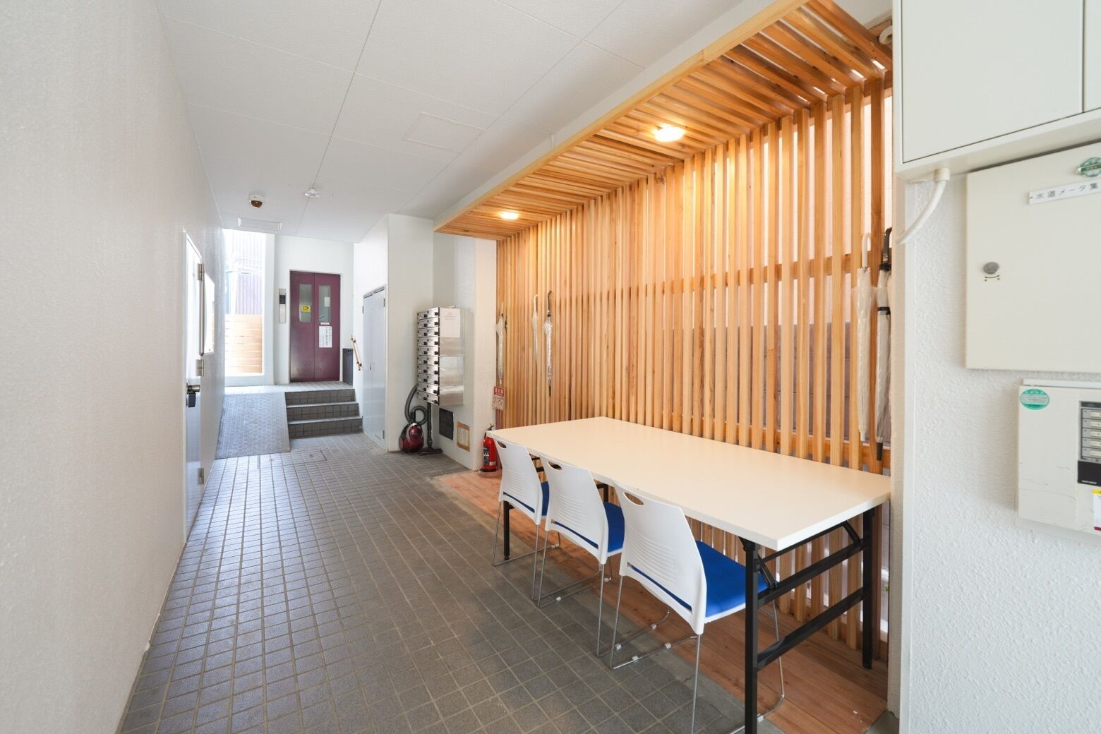 A hallway with a tiled floor, a wooden slatted wall, a white table with chairs, and a door at the end.